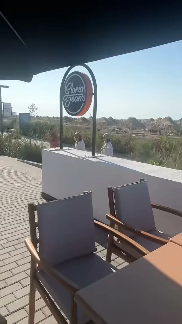 Тот же круглый знак Gloria Jean's с террасы: под чёрным зонтом, а за парапетом — пляж с соломенными зонтами и Каспий