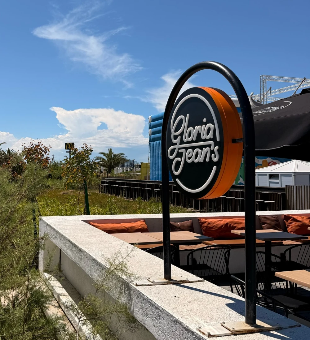 Круглый знак Gloria Jean's на дуге над террасой кофейни в Sea Breeze: чёрный круг в оранжевой обойме, под ним диваны с оранжевыми подушками; вдали видна жёлтая арка McDonald's
